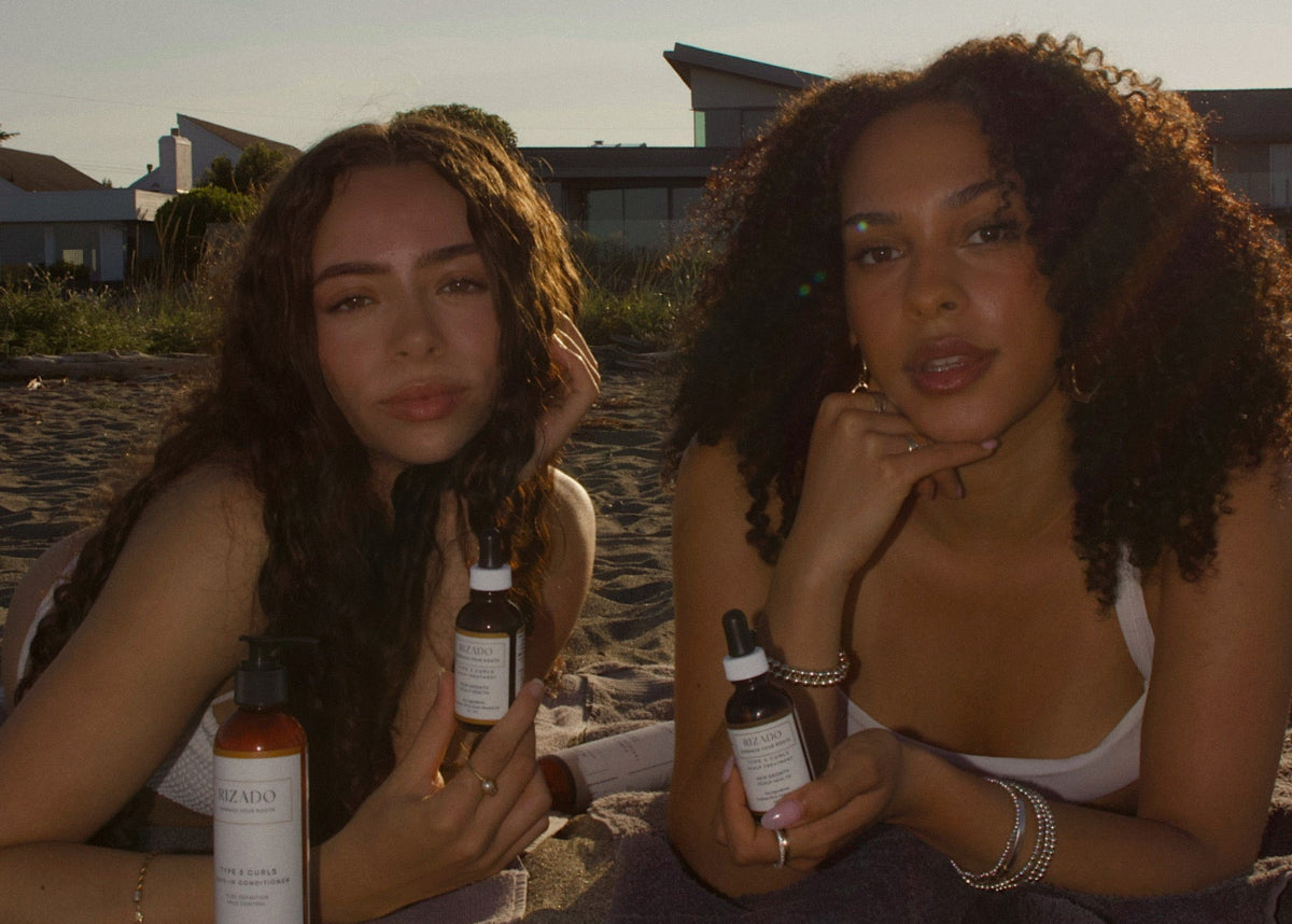 Two women sitting on a beach with bottles of a product.